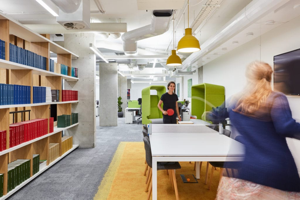 Réaménagement de l'espace de pause des bureaux avec une grande bibliothèque, un coin salon et deux employés jouant au tennis de table.